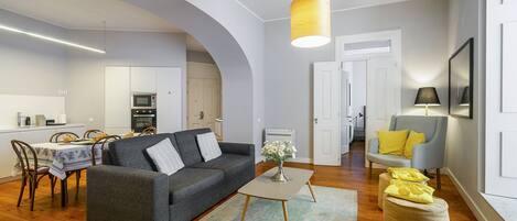 Een moderne woonkamer met een grijze bank, een salontafel en poefjes. Er is een eethoek met een tafel en stoelen, en een keuken op de achtergrond.