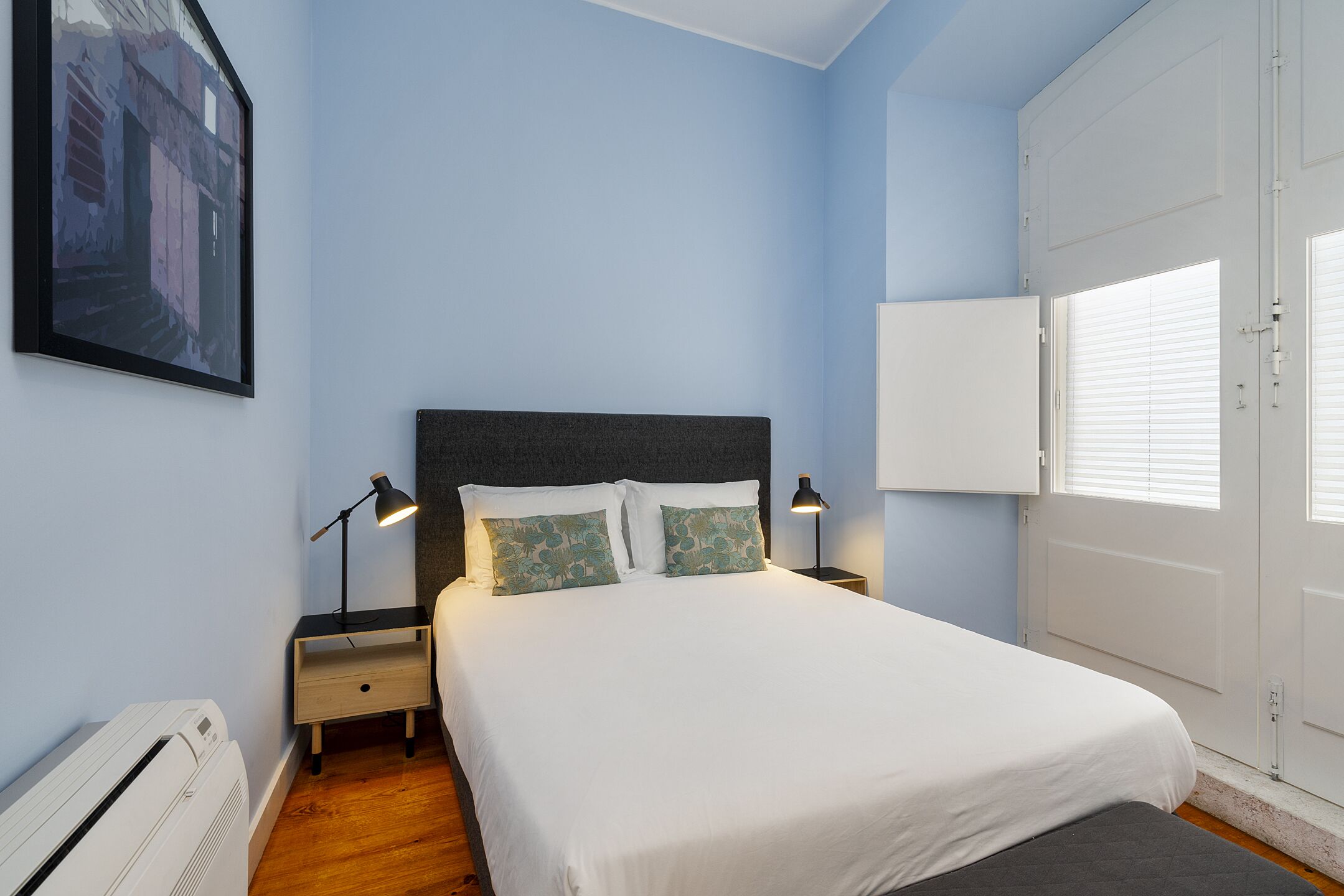 Une chambre à coucher comprenant un lit, deux lampes de chevet, une table de chevet, un radiateur et une porte avec des stores.