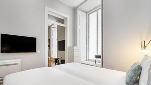 Una habitación de hotel moderna con una cama grande, un televisor de pantalla plana, una mesita de noche y un ventanal con persianas.