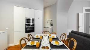 Una cocina moderna con horno y microondas integrados, una mesa de comedor con platos y vasos, y un sofá gris en el área de estar contigua.