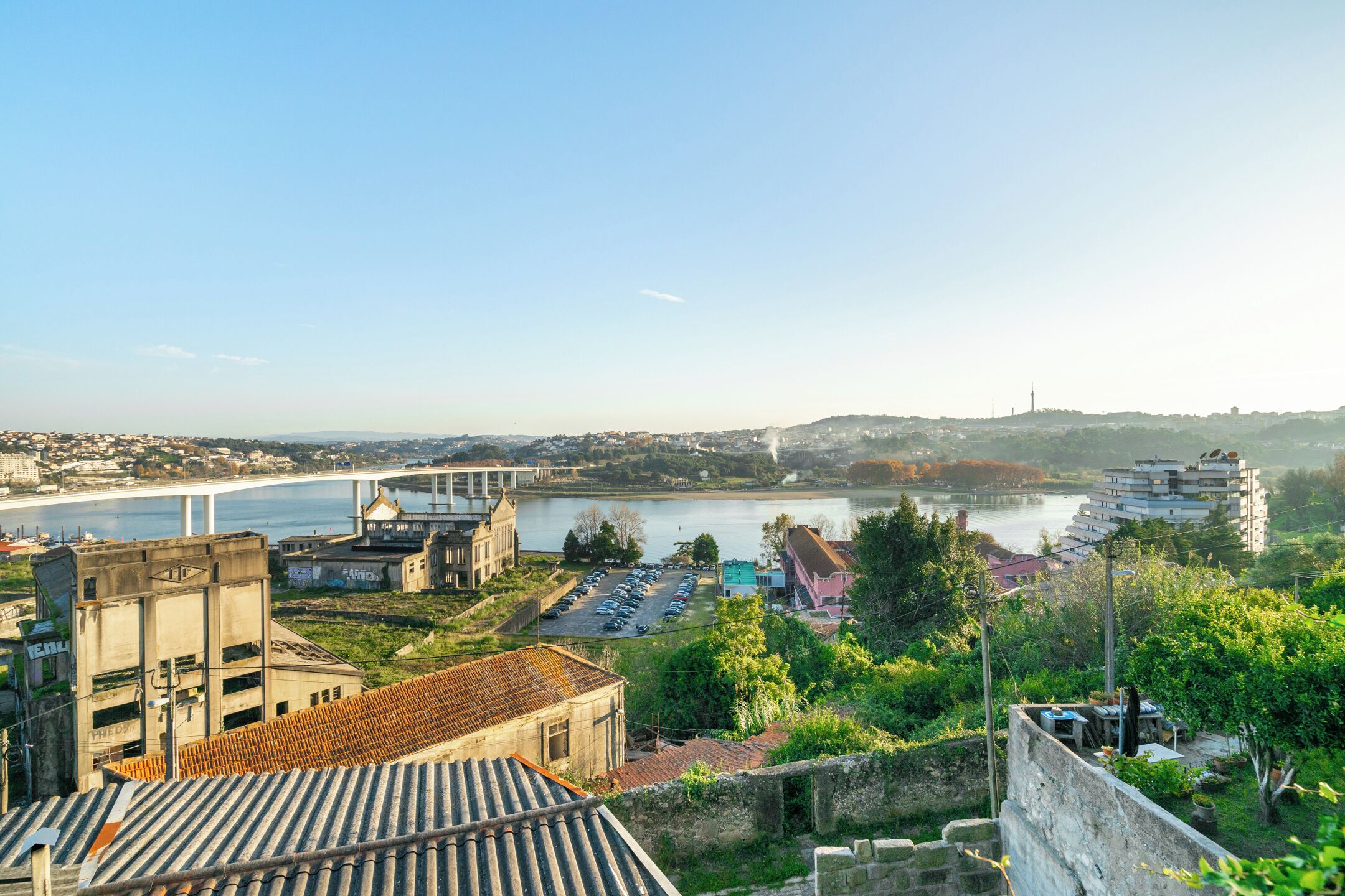 A view of a riverside cityscape with a bridge, buildings, and a parking lot.