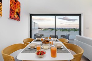 Mesa de desayuno servida, vistas de la ciudad a través de amplios ventanales y mobiliario moderno.