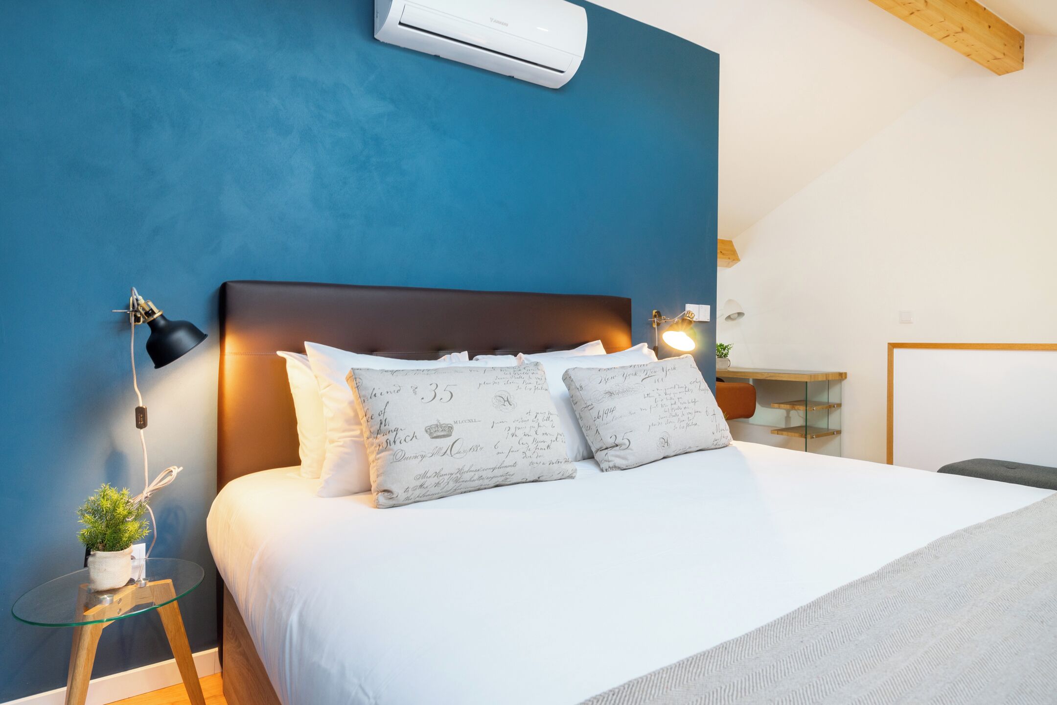 Quarto com cama, luminárias de cabeceira, uma mesinha com uma planta e uma escrivaninha com cadeira.