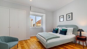 Ein Schlafzimmer mit Bett, einem Sessel, einem Kleiderschrank, einem Fenster und zwei gerahmten Bildern an der Wand.