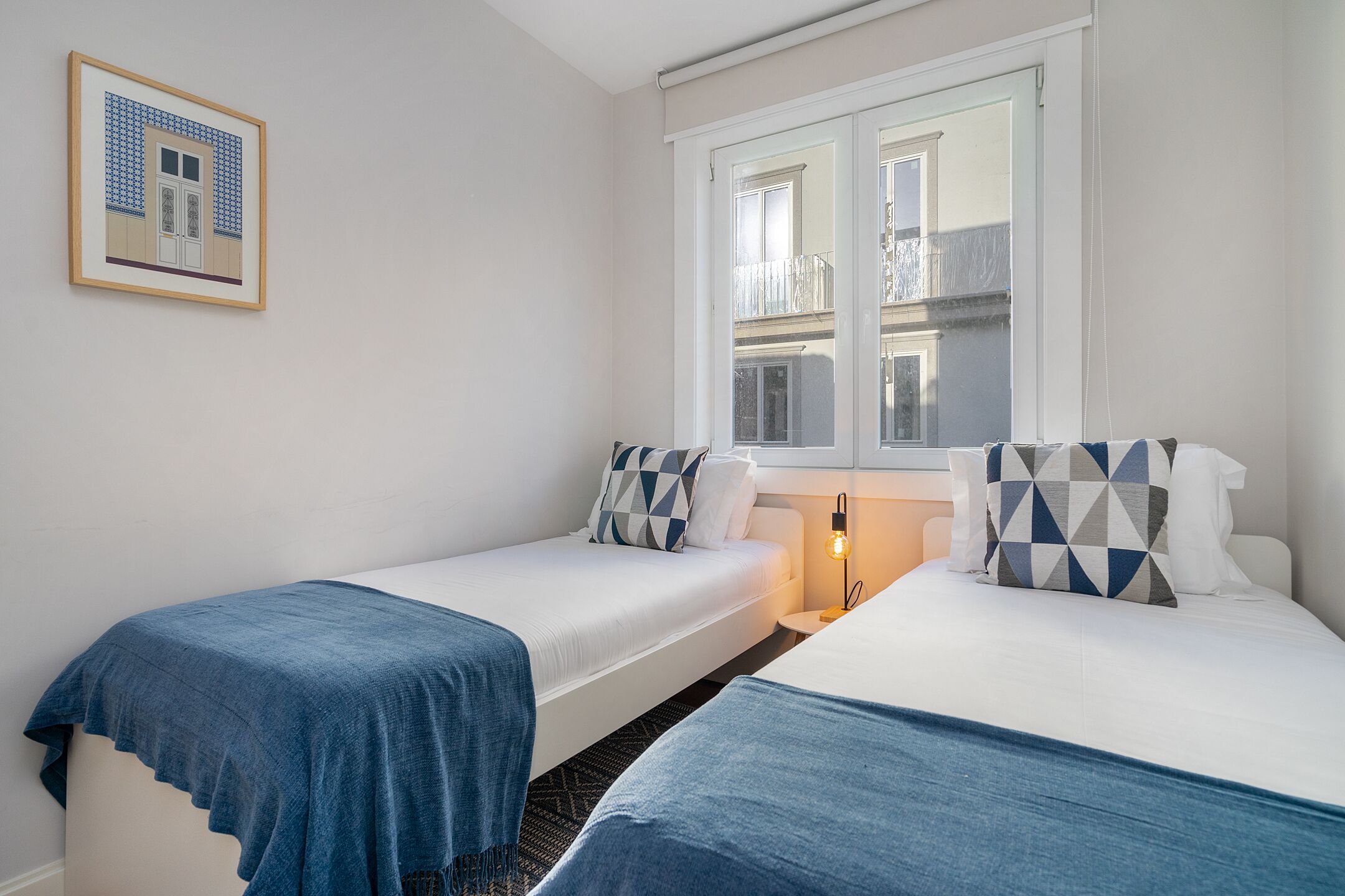 Une chambre d’hôtel avec deux lits simples, une fenêtre donnant sur l’immeuble et un tableau encadré au mur.