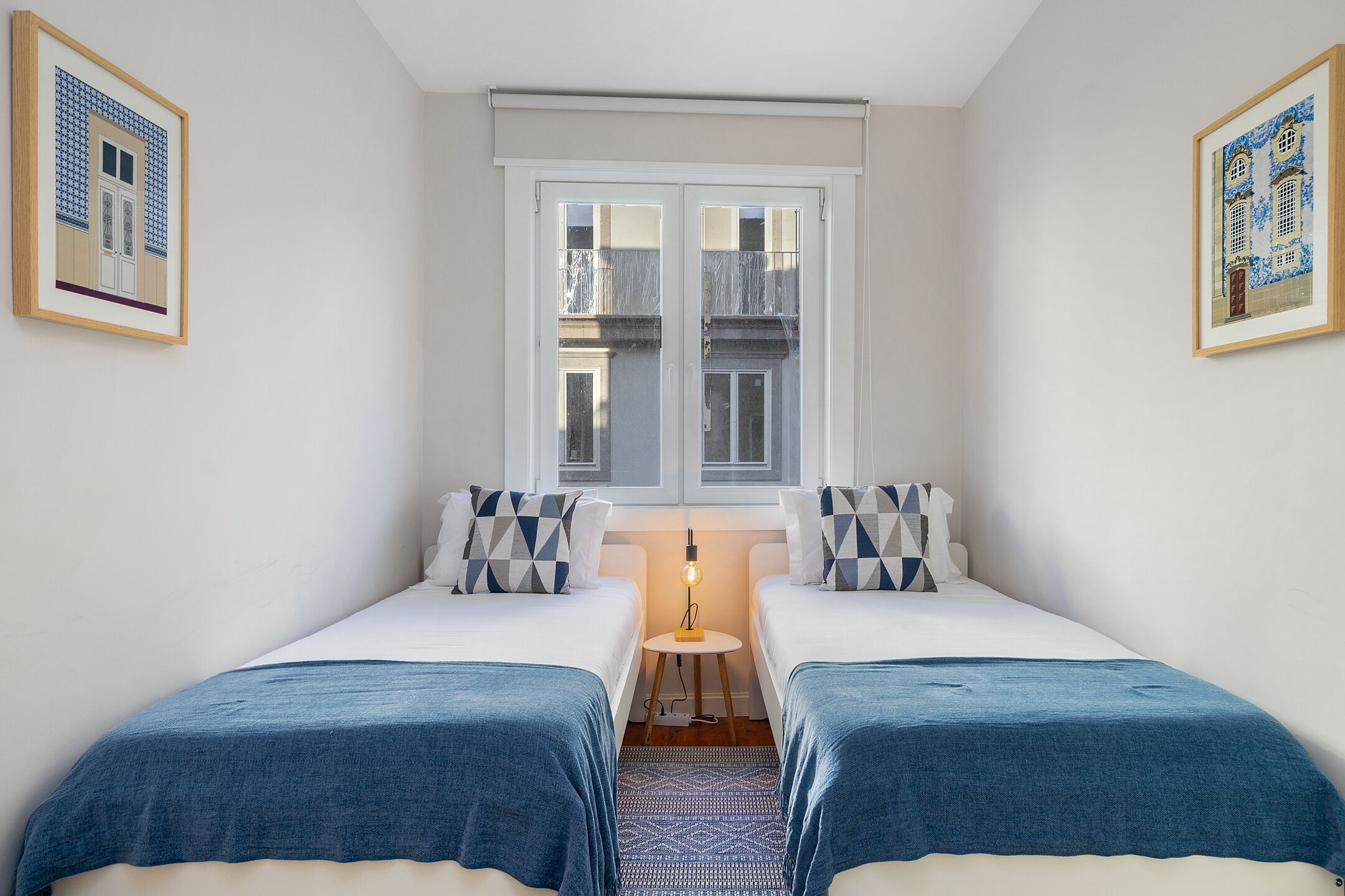 Ein Zimmer mit zwei Betten, einem kleinen Tisch mit Lampe und einem Fenster mit Blick auf Gebäude.
