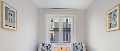 Ein Zimmer mit zwei Betten, einem kleinen Tisch mit Lampe und einem Fenster mit Blick auf Gebäude.