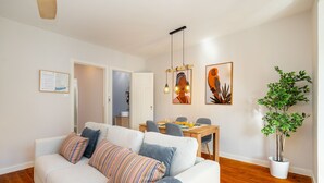 Ein Wohnzimmer mit einer Couch, einem Esstisch und Stühlen. Eine Topfpflanze und Kunstwerke zieren die Wände.