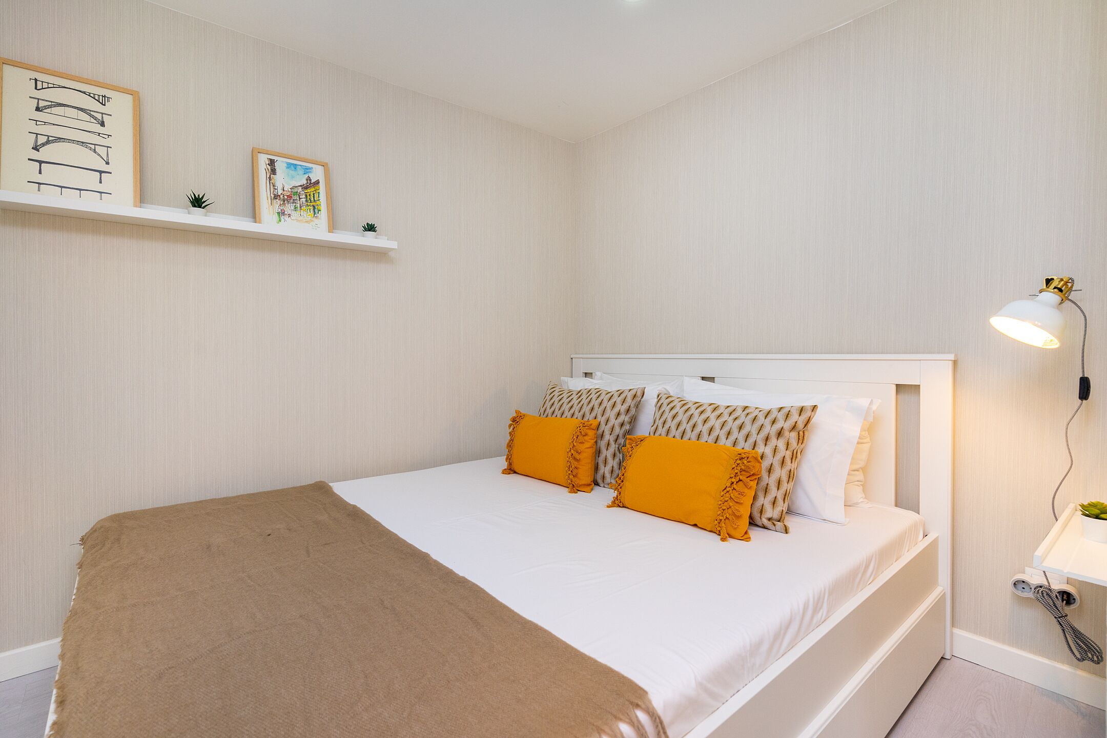 A neatly arranged bedroom with a bed, a bedside lamp, and wall-mounted shelves with framed pictures.