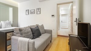 En moderne stue med sofa, tv og et lille bord. Der er en dør til et andet rum og en vægmonteret aircondition.