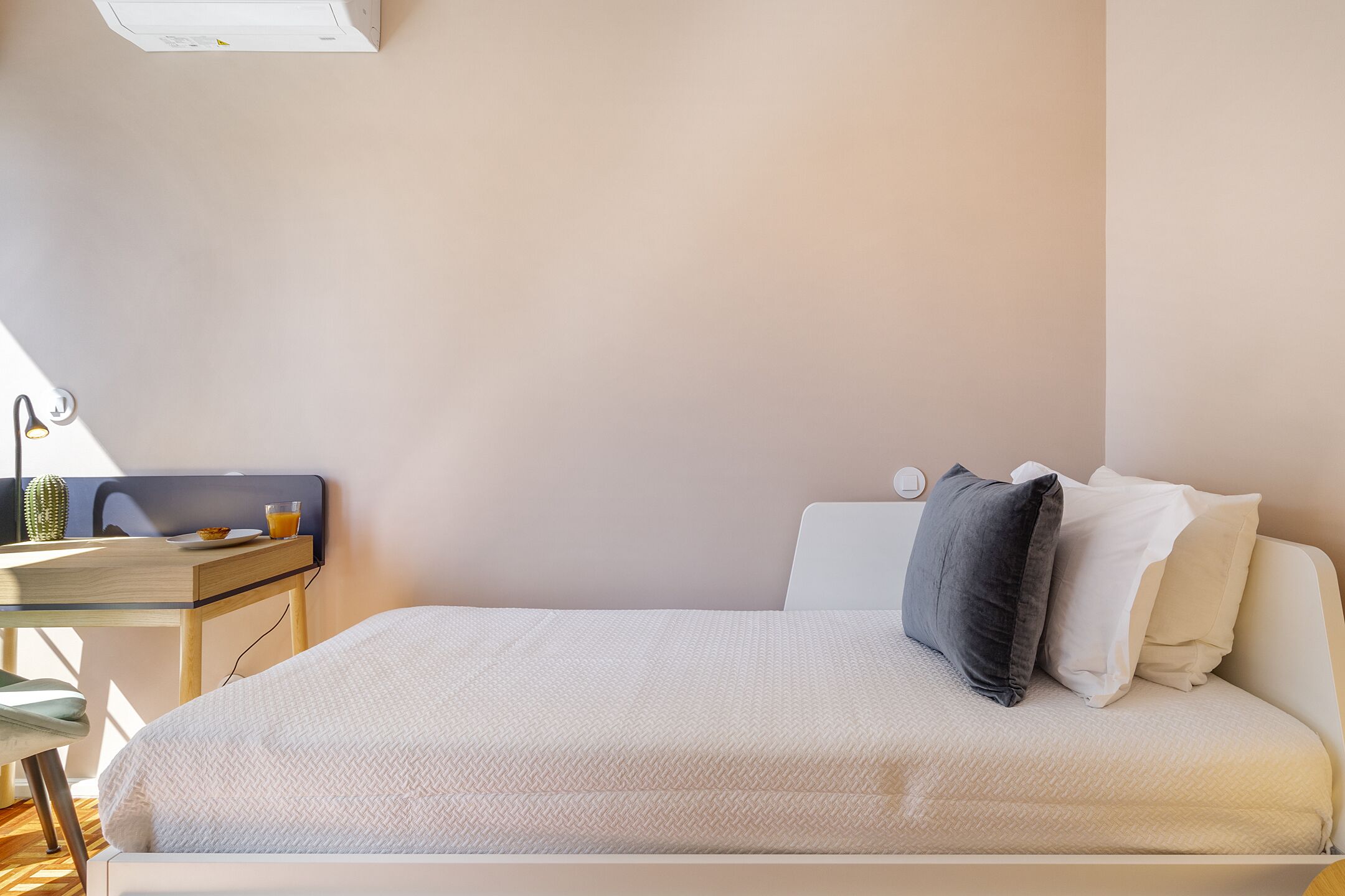 A neatly made bed with a white comforter and pillows, a small bedside table with a lamp, and a cactus on the floor.