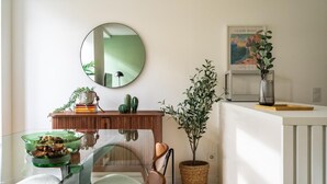 A dining area with a glass table, chairs, a round mirror, a wooden sideboard, a potted plant, and a framed artwork on the wall.