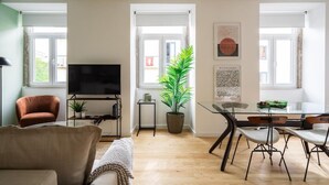 A modern living room with a flat-screen TV, a sofa, a dining table with chairs, and a potted plant.