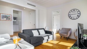 En moderne stue med en grå sofa, et spisebord med tallerkener og et glas appelsinjuice, en grå lænestol og et stort vægur.