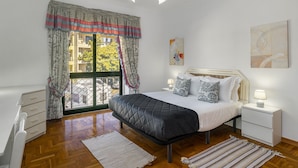 Un dormitorio con cama, mesitas de noche, cómoda, ventana con cortinas y vistas a edificios.