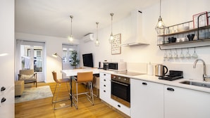 A modern kitchen with white cabinets, a built-in oven, a microwave, and a sink. There are bar stools, a dining table, and a plant in the room.