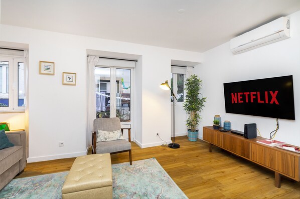 A modern living room with a large flat-screen TV displaying "NETFLIX," a wooden console table with electronic devices, a grey armchair, a beige ottoman, and a patterned rug. - Green Street 1-Bed Perfect To Explore Lisbon (Lisbon)
