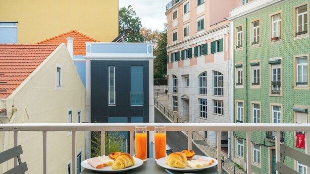 A balcony with two plates of croissants and orange juice, overlooking a street with colorful buildings.