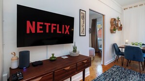 A modern living room with a large flat-screen TV mounted on the wall displaying "NETFLIX," a wooden console table with a speaker, a remote control, and a small plant.