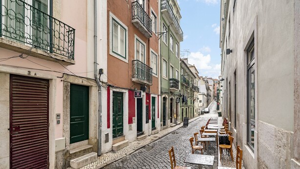 Una estrecha calle adoquinada flanqueada por edificios de colores, cada uno con una persiana cerrada y un pequeño escalón que conduce a la entrada.