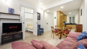 Una sala de estar moderna con una televisión de pantalla plana, un sofá, una mesa de comedor con sillas y una cocina al fondo.