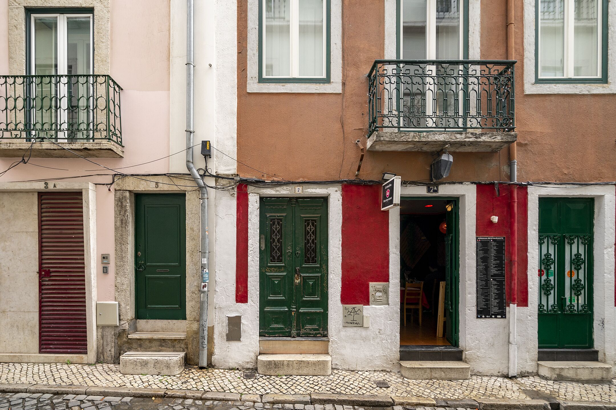 Una hilera de edificios con puertas verdes y rojas, una calle empedrada y un balcón visible con barandillas de metal.