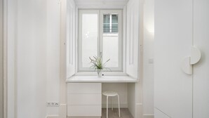 Una habitación blanca con ventana, un escritorio y un taburete.