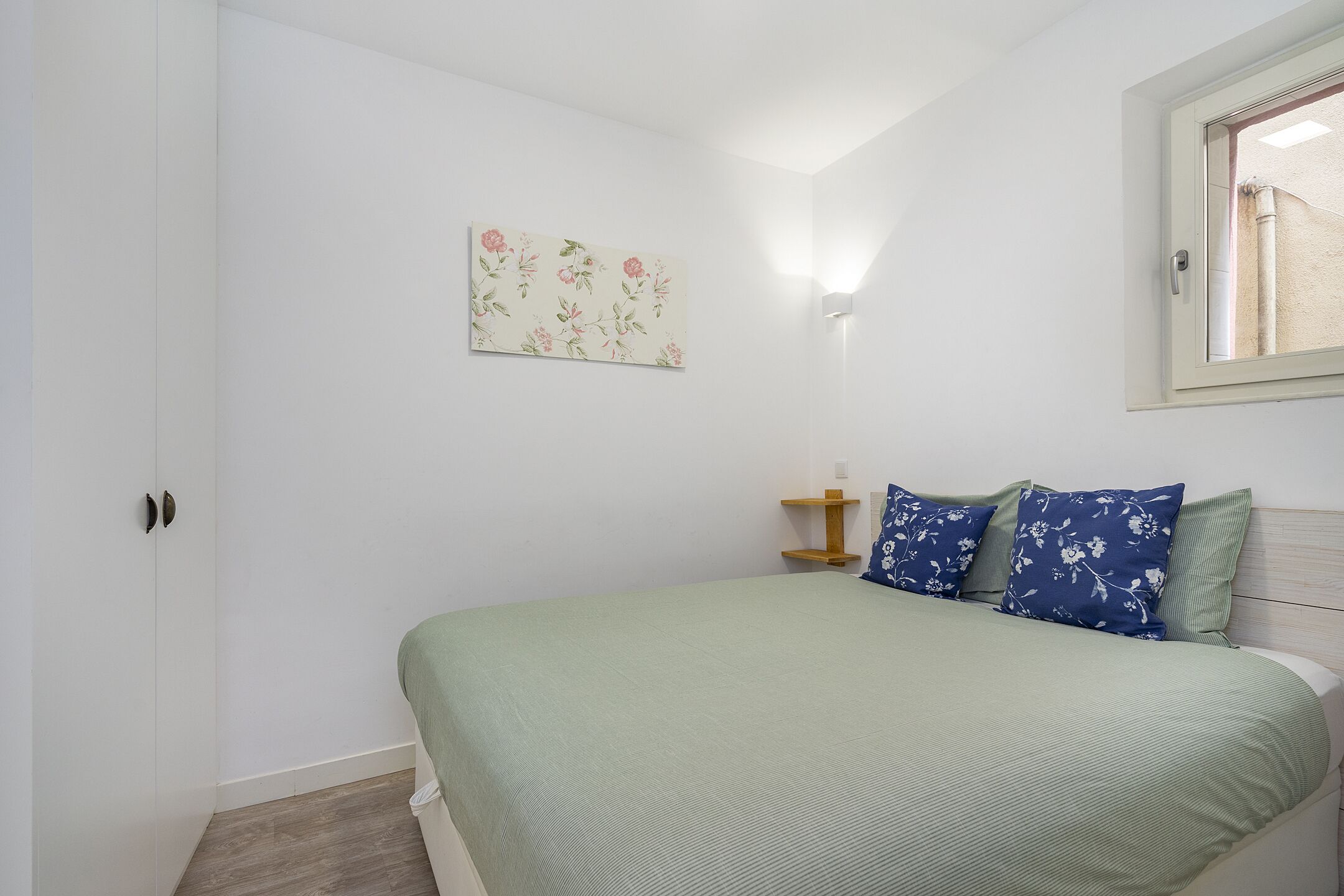 A bedroom with a bed, a nightstand, a wall-mounted light, and a window.