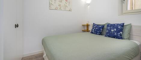A bedroom with a bed, a nightstand, a wall-mounted light, and a window.