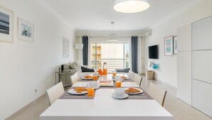 A modern dining area with a long table set for breakfast, a TV on the wall, and a balcony view. - Family Holidays W/ S-Pool, Ac, Elevator & Garage (Loulé)