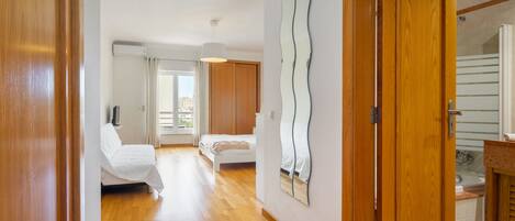 A hotel room with a wooden floor, a bed, a bathroom with a mirror, and a view of the city through the window.