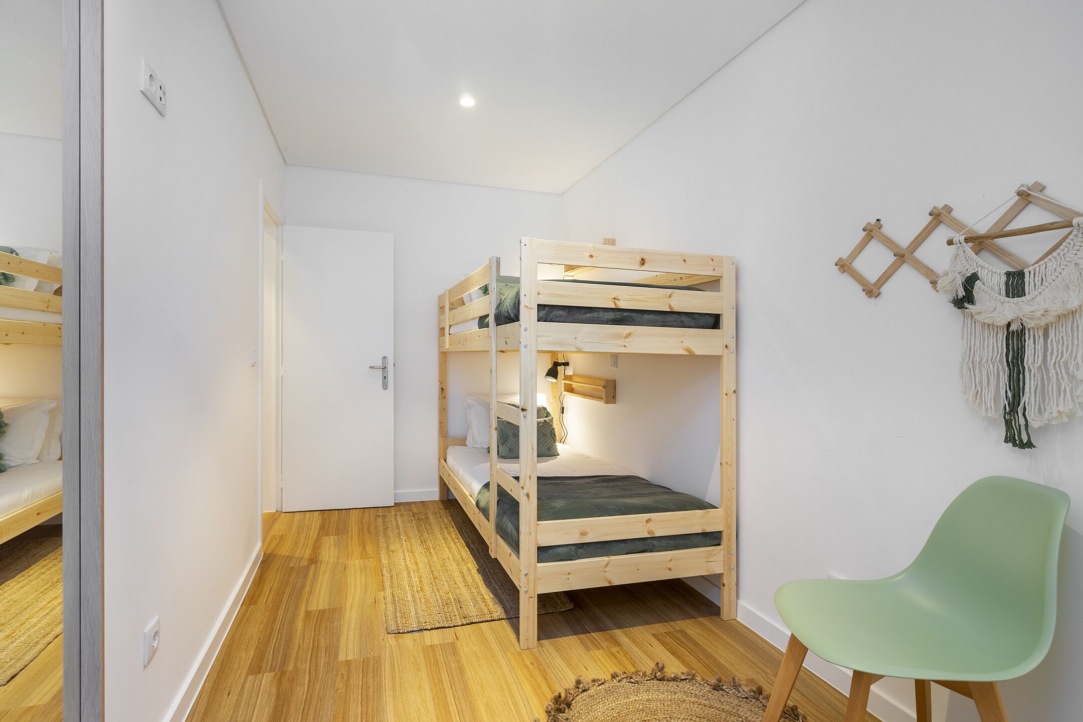 Ein Stockbett mit Holzbettgestell und einer grünen Matratze, ein hellgrüner Stuhl und ein gewebter Teppich auf Holzdielen.