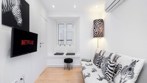 Un salon moderne avec un canapé blanc, des coussins à motifs zébrés, un téléviseur fixé au mur qui affiche « NETFLIX », une lampe murale à motifs zébrés et une fenêtre avec des stores.
