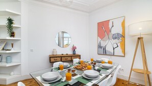 Un coin repas avec une table en verre, des chaises blanches et une table dressée pour le petit-déjeuner, comprenant des assiettes, des verres et du jus d’orange.