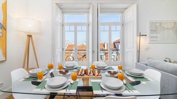 Un coin repas avec une table en verre, des chaises blanches et un assortiment de produits pour le petit-déjeuner, notamment des croissants et du jus d’orange.