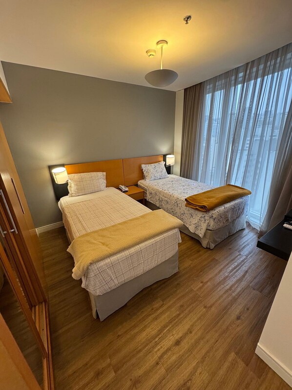 Habitación de hotel con dos camas, suelo de madera, un ventanal grande con cortinas e iluminación en el techo.