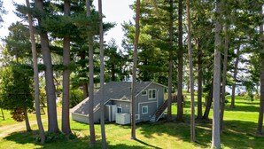 Une petite maison entourée de grands arbres, avec une cour verdoyante et un escalier menant à l’entrée.
