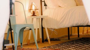 Ein Zeltraum mit einem Metallbettgestell, einem kleinen Tisch mit Lampe und einem blauen Stuhl.