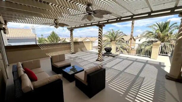 Un patio al aire libre con ventilador de techo, asientos acolchados y vistas a palmeras y edificios.
