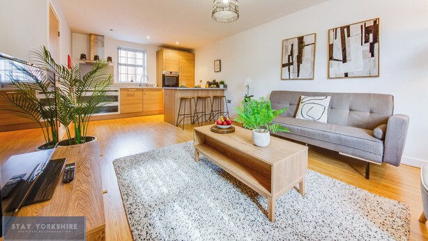 Una sala de estar moderna con un sofá gris, una mesa de centro de madera y una cocina al fondo.
