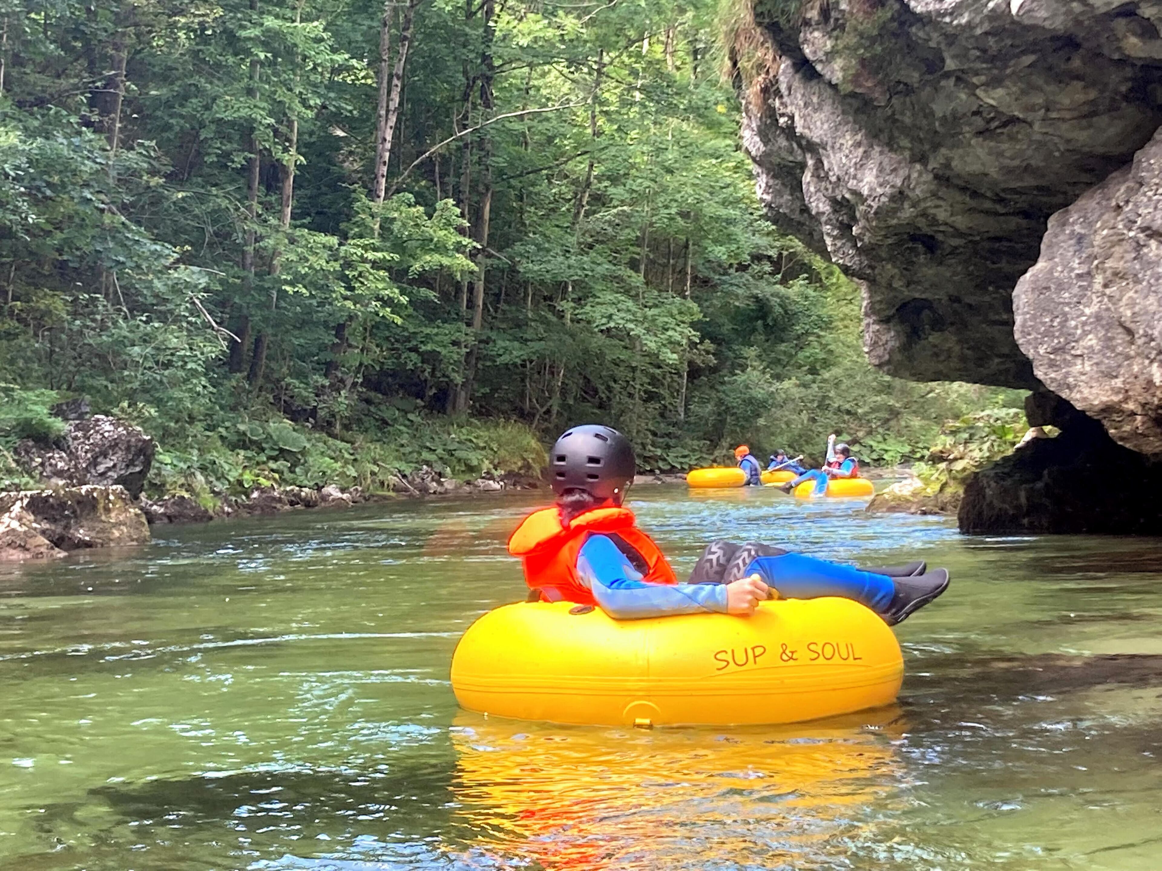 A person in a yellow inflatable tube with "SUP & SOUL" written on it, floating on a river surrounded by greenery and rocks.
