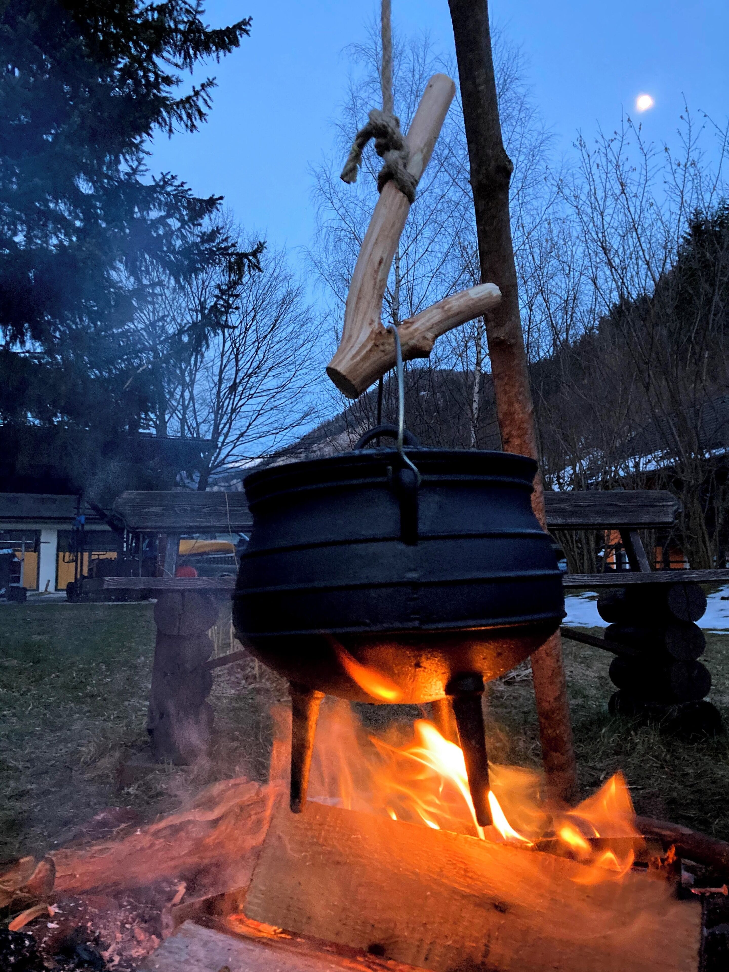 A portable cooking setup with a pot suspended over an open fire, surrounded by trees and a building in the background.