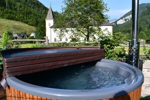 Un bain à remous avec une structure extérieure en bois et un couvercle en métal, situé dans un jardin, avec un bâtiment blanc et une église en arrière-plan.