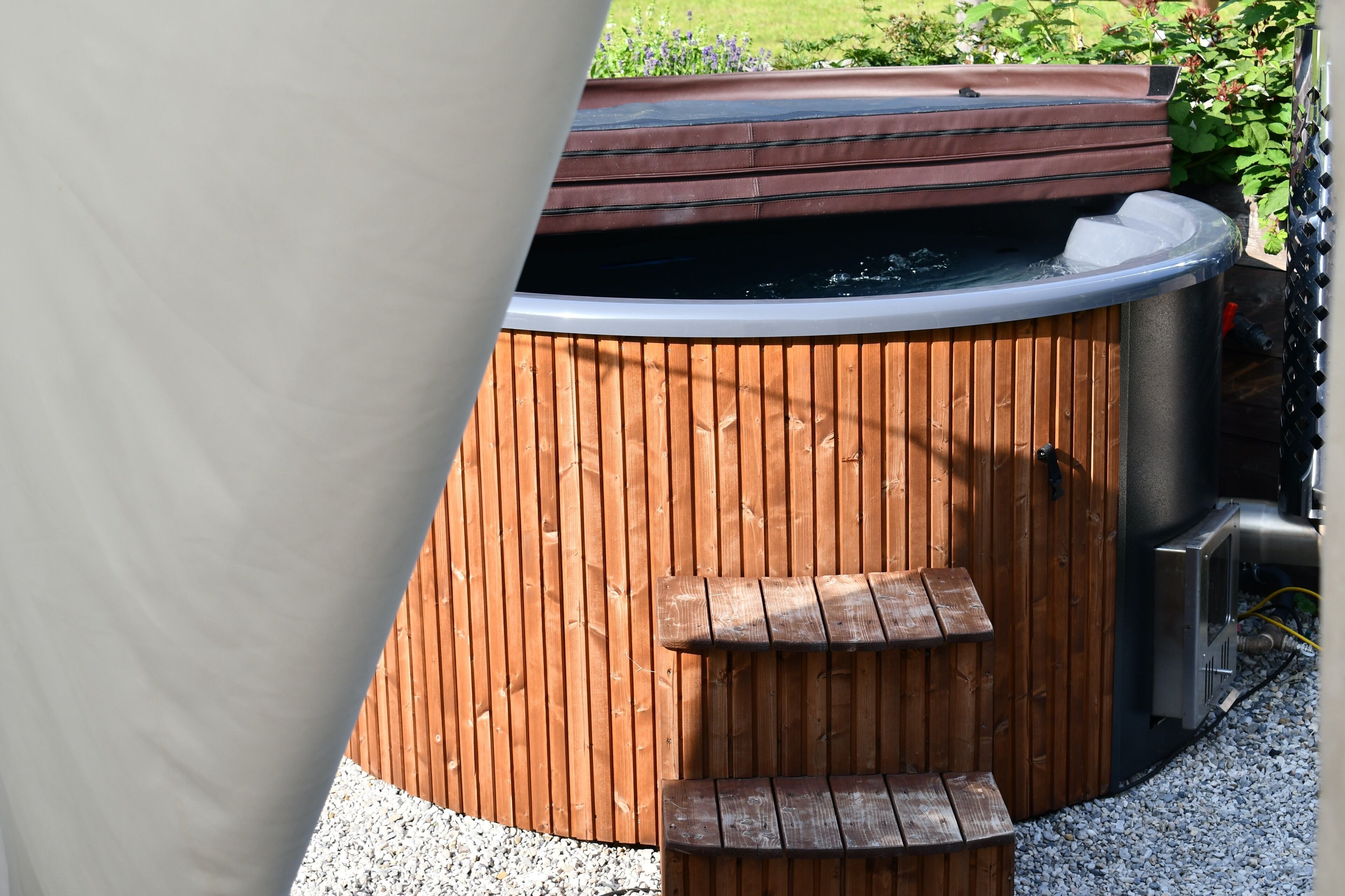 A hot tub with a wooden exterior and a metal frame, surrounded by gravel and plants.