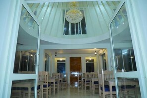 A dining area with a high ceiling, a chandelier, and a view through large windows. - Srinivasa Hotel (Chittoor District)