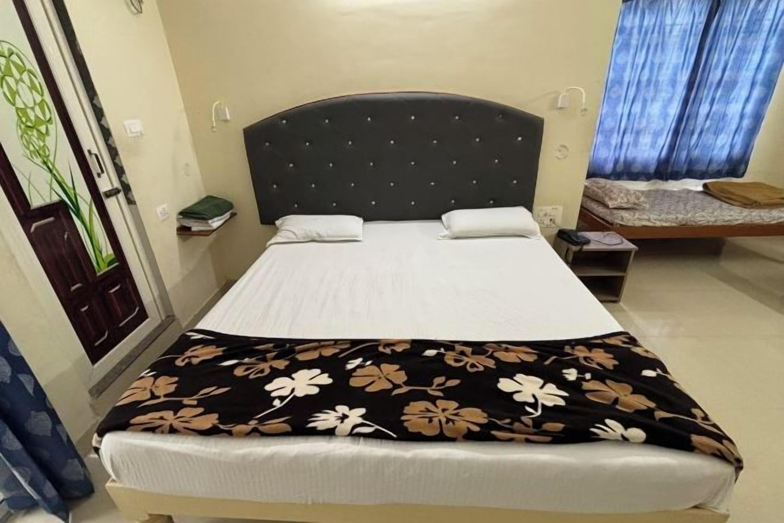 A hotel room with a bed, a bedside table, a nightstand, a window with curtains, and a door with a decorative panel.