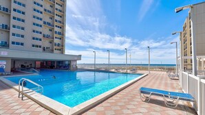 Piscina com vista para o oceano, rodeada por espreguiçadeiras e um edifício com vários andares.