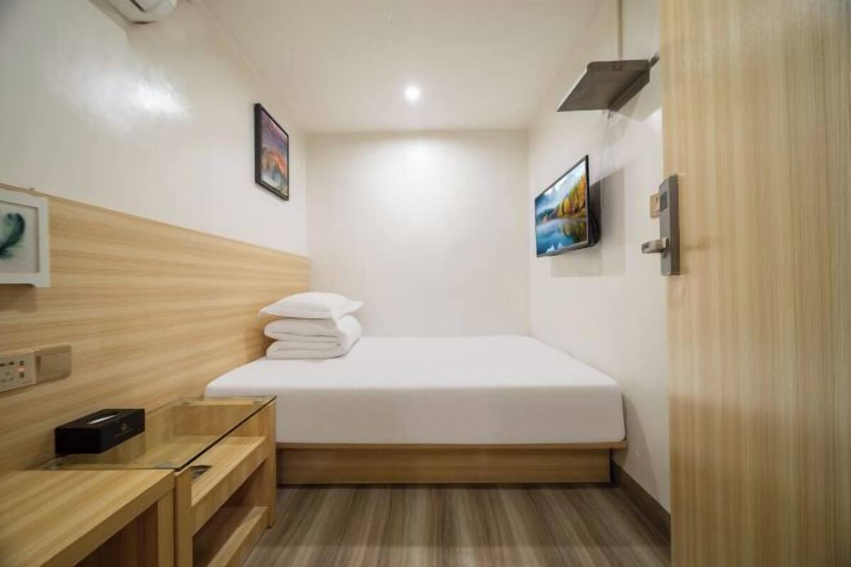 Una habitación de hotel pequeña y ordenada con una cama individual, una mesita de noche, un televisor montado en la pared y un panel de pared de madera.