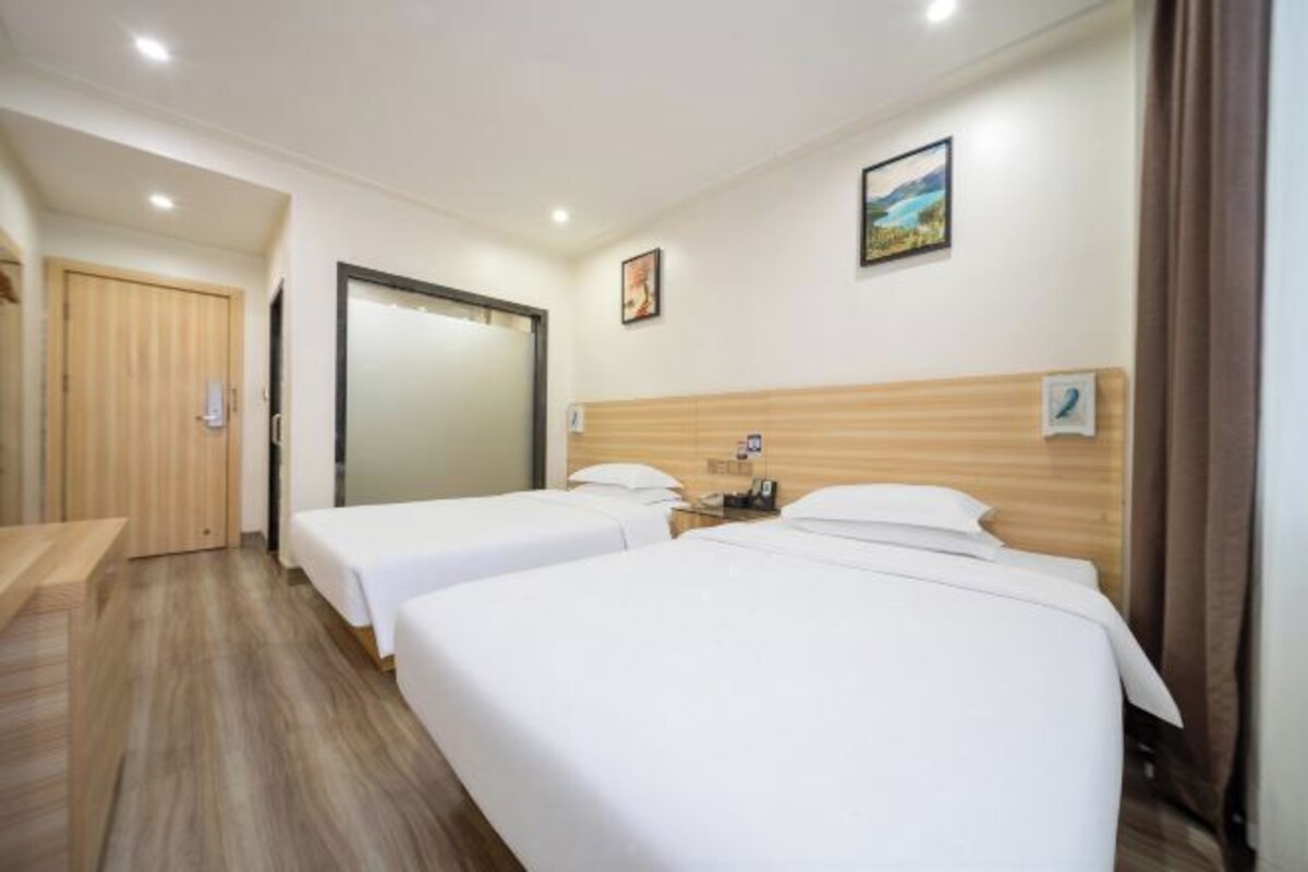 Ein Hotelzimmer mit zwei Einzelbetten, einem hölzernen Kopfteil, einem kleinen Nachttisch, einer an der Wand montierten Uhr und einer Tür zu einem anderen Raum.