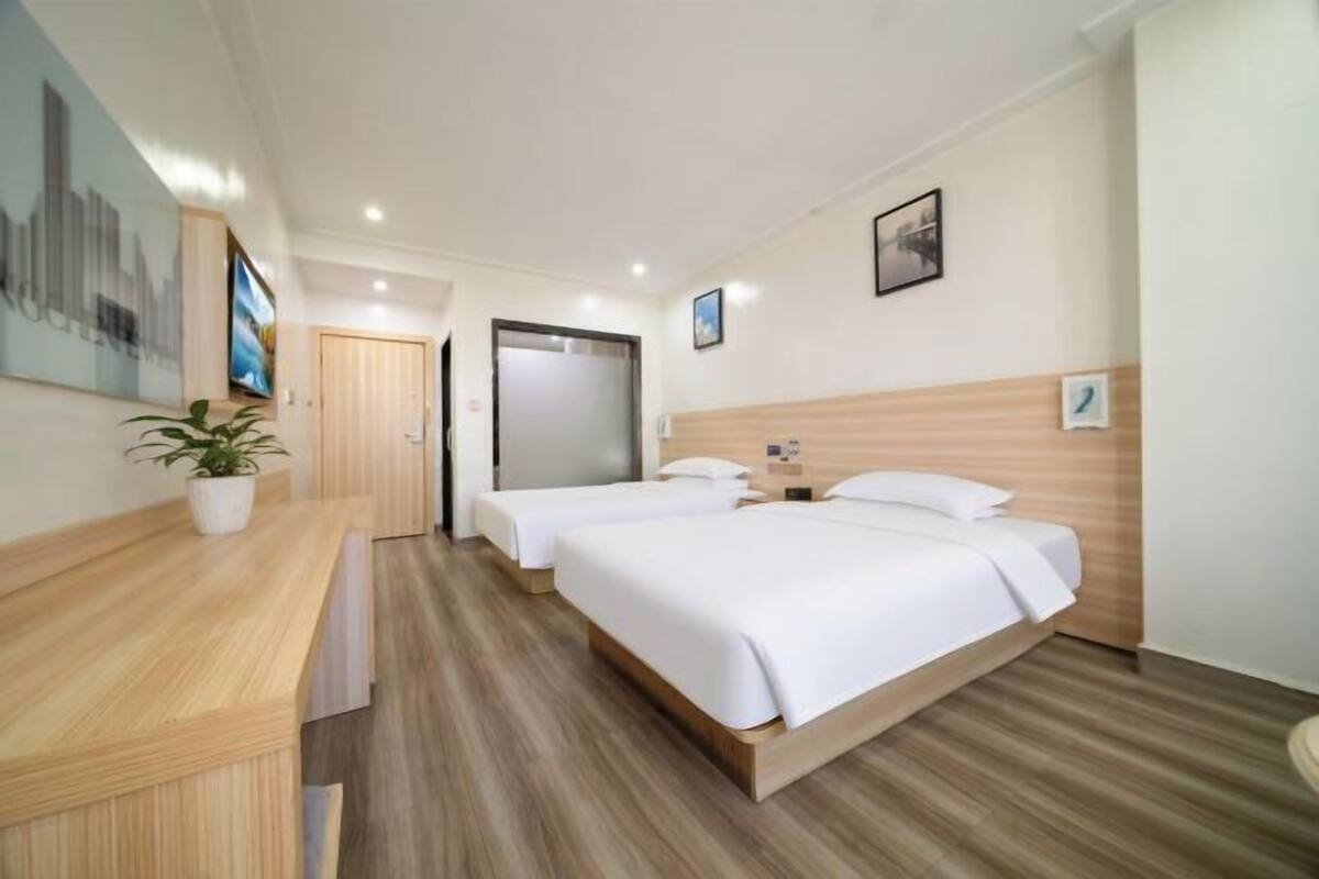 Habitación de hotel con dos camas, un escritorio de madera, una planta en maceta y una pintura con vista a la ciudad.